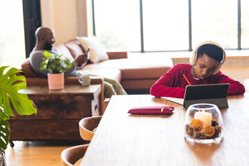 African American father using smartphone, son working on tablet at dining table, copy space