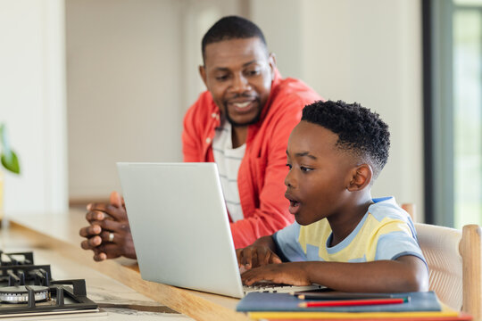African American father and son leaning over home kitchen island using laptop and colored pencils