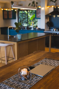 Glass bowl holding lit candle and pinecones is glowing on table near laptop in home kitchen