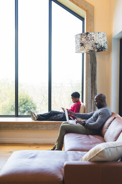 African American father and son at home, son on bench using smartphone, father typing on laptop