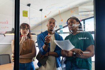 Diverse coworkers brainstorming strategy chart in office on glass whiteboard using markers, tablet