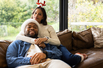 Diverse couple hugging on brown leather sofa by large window wearing antler headband, Santa hat