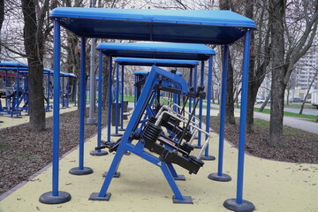 row of blue outdoor gym machines with protective canopies on rubberized surface in city park for public fitness and active longevity programs