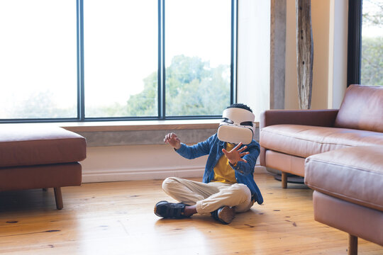 African American boy exploring virtual reality headset while sitting on wooden floor in living room