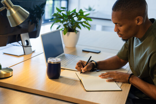 Adult African American man writing in open notebook at wooden desk with laptop, monitor, copy space