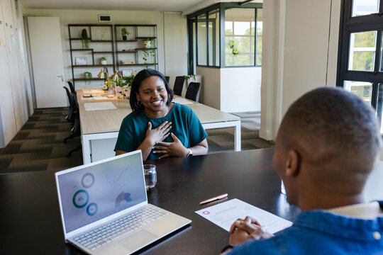 African coworkers sitting at office, woman smiling holding hand to chest, laptop showing charts