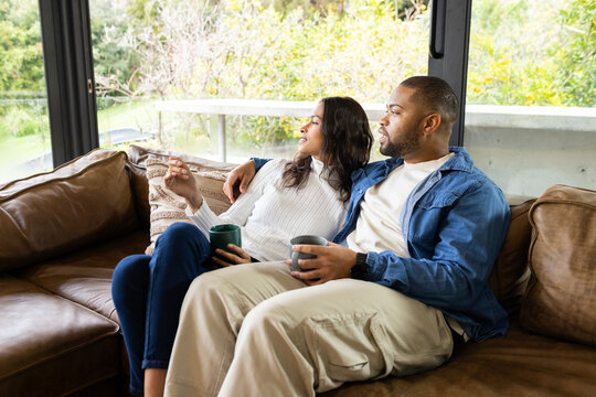 Diverse couple sitting on brown leather sofa in living room with throw pillow, holding ceramic mugs - Powered by Adobe