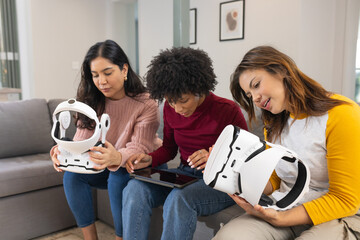 Diverse female friends exploring virtual reality headsets and tapping tablet on sofa in living room