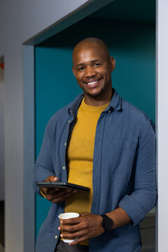 African American man standing in office hall holding tablet and coffee cup wearing sweater