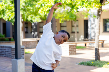 African boy stretching sideways at courtyard near brick planters, wearing white tee and dark shorts