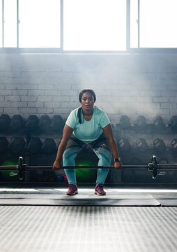 Mid adult African American woman lifting loaded barbell from mat at gym, wearing teal outfit