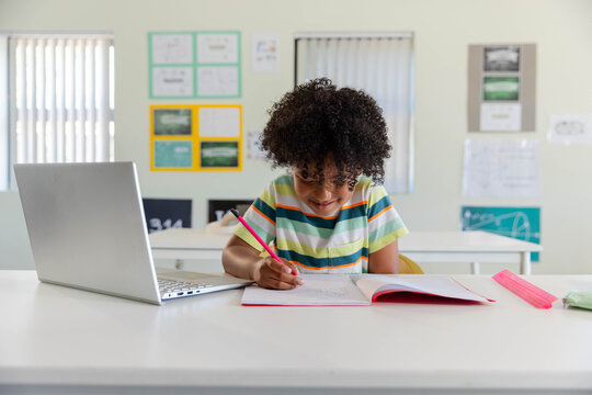 African American youth in striped tee sitting at classroom desk writing in pink notebook by laptop