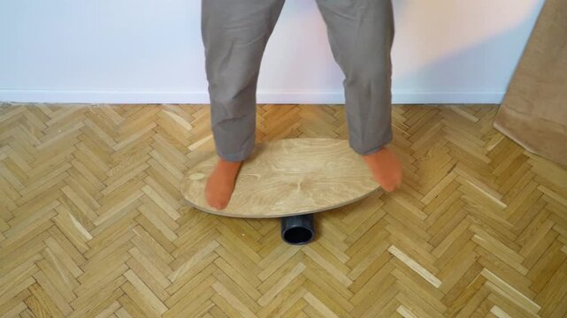 lowmotion barefoot person in casual pants balancing on wooden balance board with foam roller underneath training vestibular system and core stability indoors