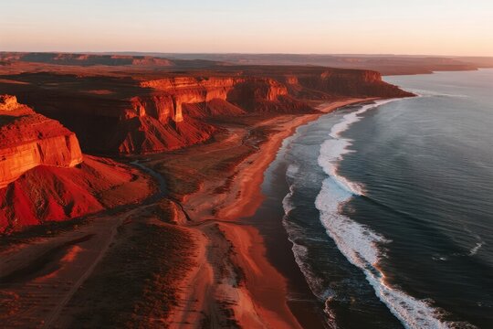Aerial view of a dramatic coastal landscape at sunset with red rock formations and waves crashing along the shore - Powered by Adobe