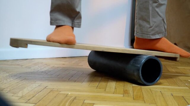 lowmotion barefoot person in casual pants balancing on wooden balance board with foam roller underneath training vestibular system and core stability indoors