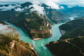 Aerial view of a turquoise river winding through mountainous terrain with clouds hovering over peaks