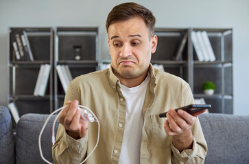 Upset frustrated man holding broken mobile phone and charger with cable in hands. Charging failure...