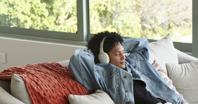 Man glancing, reaching for white headphones, adjusting and reclining on sofa beside window to relax
