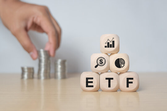 wooden cube with ETF text , Exchange Traded Fund and blurred hand with stack of coins. for business finance conceptual