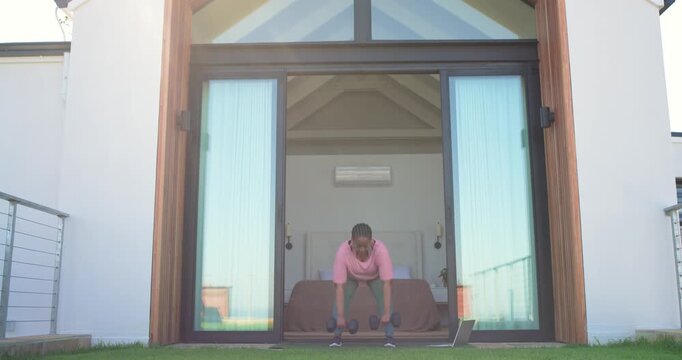 African American woman doing sumo squats with dumbbells in backyard while following laptop workout