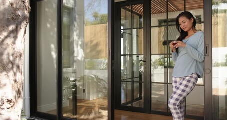 Asian woman entering through sliding door scrolling smartphone messages gazing at plants copy space - Powered by Adobe