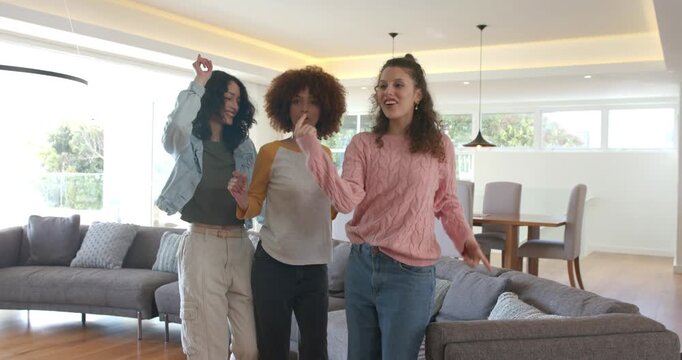 Diverse female friends raising fingers in living room guiding friend onto sofa dancing for fun