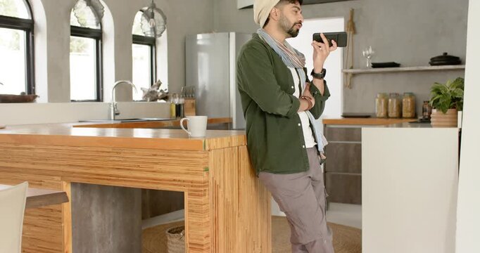 Indian man leaning against kitchen island speaking into phone fidgeting near coffee mug, copy space