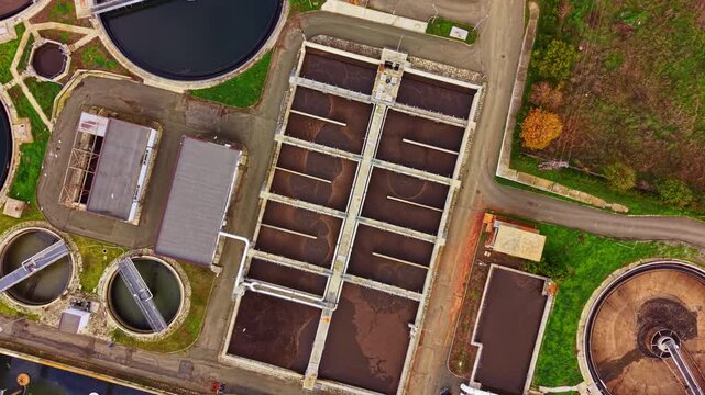 Aerial view showcases the organized structure of a wastewater treatment plant with round tanks and rectangular basins surrounded by greenery and sealed buildings on a clear day.