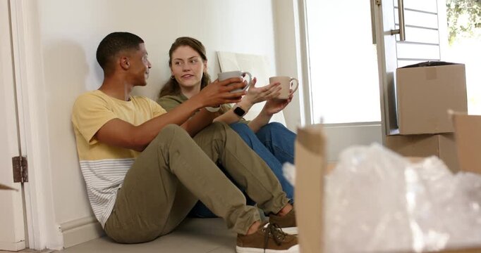 Diverse couple sitting on floor by moving boxes, taking break from unpacking, sipping coffee mugs