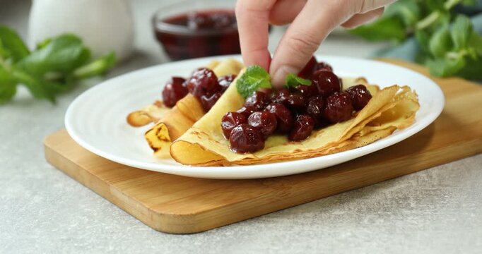 Woman adding mint to tasty crepes with cherries at grey table, closeup