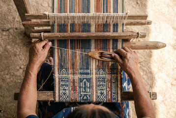 High-angle close-up of a weaver's hands passing a wooden shuttle with blue and orange thread through the warp of a traditional wooden loom to create patterned textile.