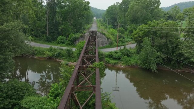 An abandoned railway bridge stretches over a calm river. Dense trees and vegetation cover the area, creating a serene and peaceful atmosphere.