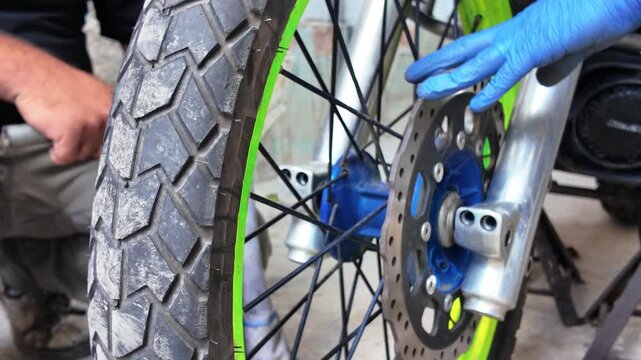 Close-up mechanics servicing motorcycle front wheel. One person holds wheel assembly while another stabilizes brake disc. Tire tread, spokes, brake rotor, and front suspension during maintenance