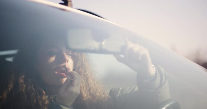 A young woman wearing braces is captured applying lipstick in a car, using the rearview mirror for assistance, reflecting a moment of everyday beauty rituals.