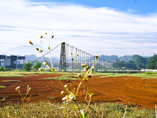 A landscape view featuring mountains, a suspension bridge, and red earth in the foreground with blue clouds in the distance. A natural morning view.