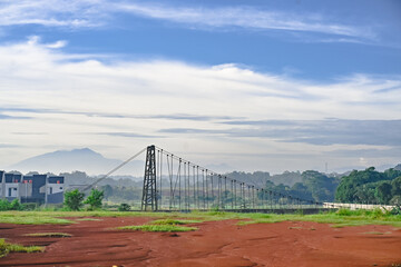A landscape view featuring mountains, a suspension bridge, and red earth in the foreground with blue clouds in the distance. A natural morning view.