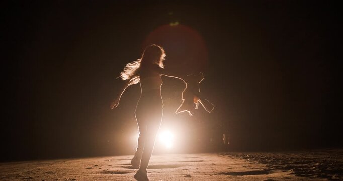 Girl throws hoodie in air while she runs towards car headlights on a dark road at night while creating a captivating silhouette