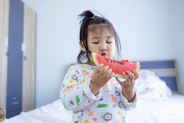 A cute little Asian girl is biting a watermelon,young girl eating watermelon against real kitchen...