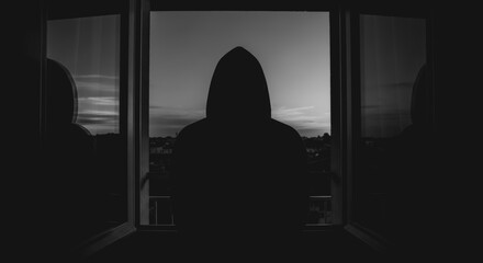 A person in a hooded sweatshirt stands by a window, gazing out at the twilight, creating a mood of solitude and introspection.