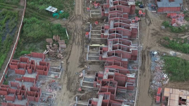 Drone footage captures an expansive Colombian construction site with red brick frames, muddy pathways, and scattered greenery, framed under overcast skies, highlighting active development.