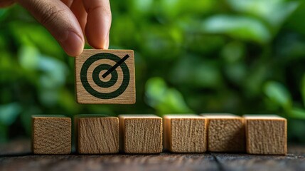 Hand placing wooden block with bullseye target symbol among plain wooden blocks on wooden surface with green blurred background symbolizing goal achievement and focus