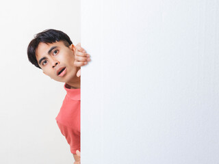 Surprised young man peeking from behind a blank wall with wide copy space, showing a humorous shocked expression suitable for ads, announcements, alerts, and creative design layouts.
