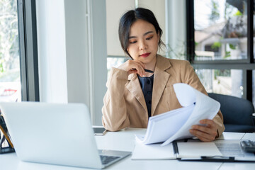 Businesswoman sitting at desk on couch in workplace or at home working on laptop and analyzing data on charts and graphs and writing on papers to make business plan and strategies for company.