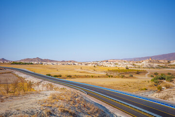 A wide landscape photograph of a scenic highway cutting through the golden plains of Cappadocia, Turkey. The scene features rolling hills, unique rock formations, and distant mountains