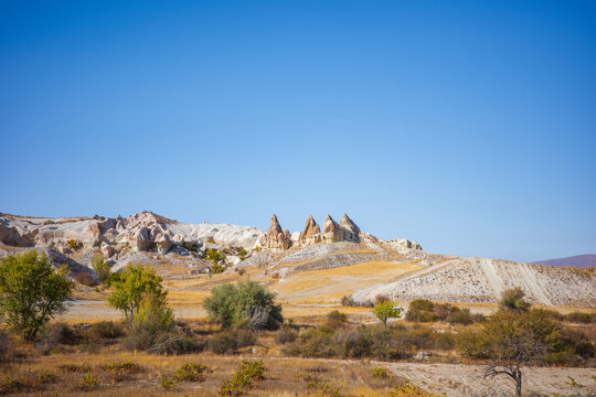 A wide-angle landscape photograph of Cappadocia, Turkey, showcasing the region’s famous fairy-chimney rock formations, rolling golden hills, and vibrant autumn vegetation beneath a clear blue sky - Powered by Adobe
