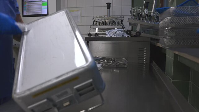 A healthcare worker wearing blue gloves handles a sterilization container with surgical instruments in a hospital reprocessing unit, ensuring hygiene and safety in a sterile medical environment.