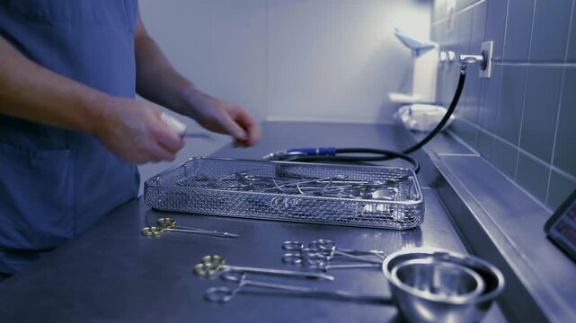 A healthcare worker in blue scrubs inspects and organizes surgical instruments in a sterilization tray inside a hospital reprocessing unit, ensuring cleanliness and patient safety.