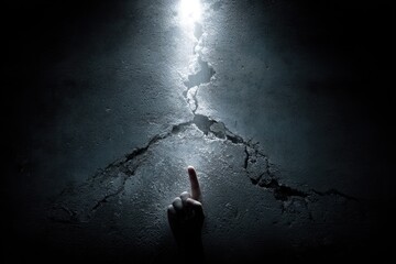A finger points at a bright light shining through a large crack in a dark, textured wall