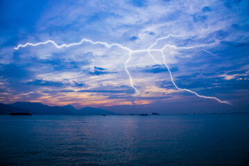A dramatic twilight seascape with vivid lightning bolts illuminating the sky above a calm ocean, with distant boats and mountains.