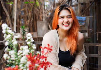 Asian happy beautiful cheerful outdoor portrait woman light orange hair smiling warmly among blooming flowers, Natural wooden textures greenery create cozy rustic garden joyful fresh nature concept.